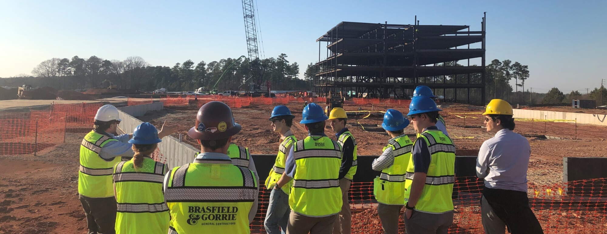 Approximately a dozen students observe a construction site from afar in hard hats and neon safety vests.