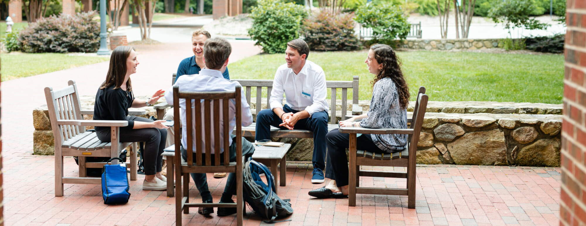 Students sit outside talking to each other. They are surrounded by green trees and lush landscape. The students are sitting on wooden outdoor furniture.