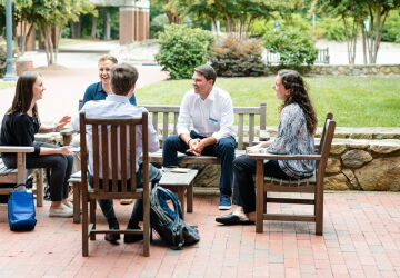 Students sit outside talking to each other. They are surrounded by green trees and lush landscape. The students are sitting on wooden outdoor furniture.