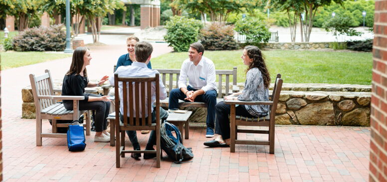 Students sit outside talking to each other. They are surrounded by green trees and lush landscape. The students are sitting on wooden outdoor furniture.