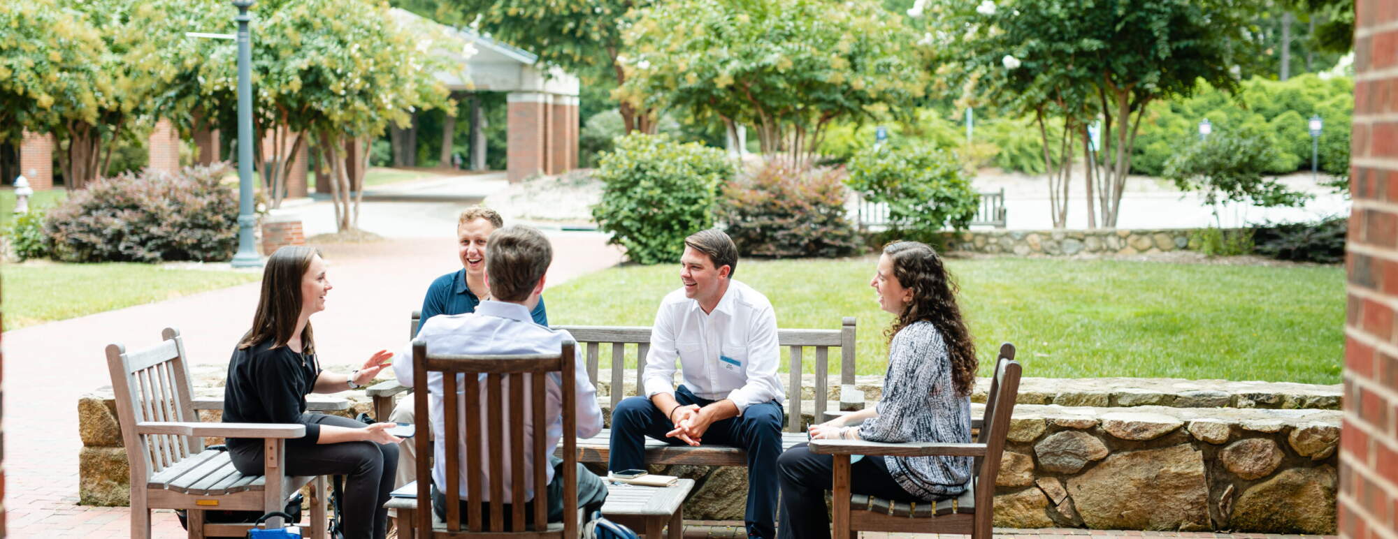 Students sit outside talking to each other. They are surrounded by green trees and lush landscape. The students are sitting on wooden outdoor furniture.