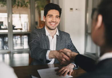 A young man in a gray blazer shakes hands with another person in an office setting.