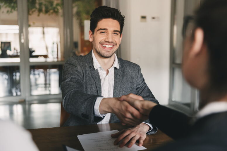 A young man in a gray blazer shakes hands with another person in an office setting.