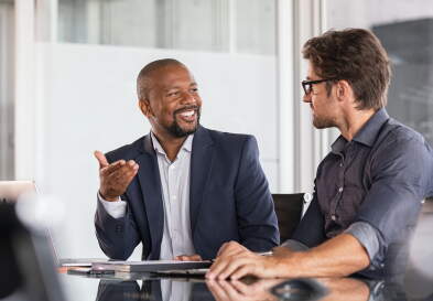 Two men in discussion at a table in an office setting. One man is wearing a navy blue blazer and smiling while gesturing; the other is wearing a dark blue shirt and glasses, listening attentively.