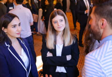 Two women look with interest at a man who is talking to them. The room behind them is full of other students in business wear - this is a networking event.