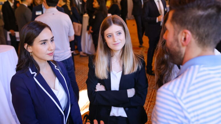 Two women look with interest at a man who is talking to them. The room behind them is full of other students in business wear - this is a networking event.