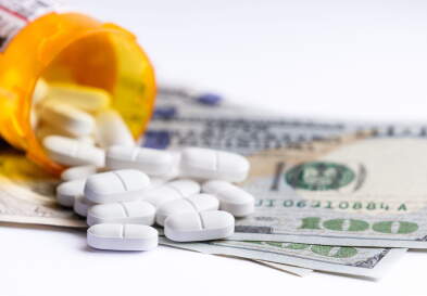 Close up of a prescription bottle with white tablets falling out on top of a group of hundred dollar bills.