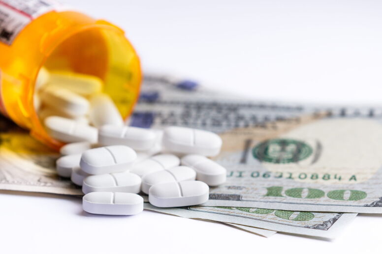 Close up of a prescription bottle with white tablets falling out on top of a group of hundred dollar bills.