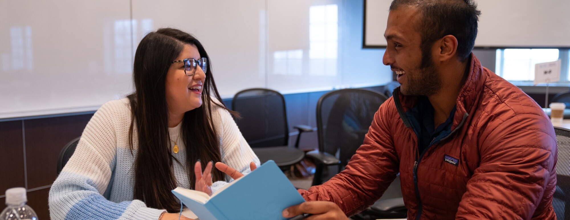 Two students are sitting at a table talking. One student is holing a notebook - it looks like they are collaborating on a project.