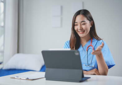 A smiling young nurse makes a video call on a tablet, gesturing with her hand while sitting at a desk.