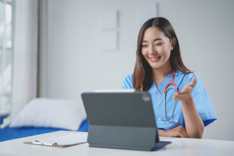 A smiling young nurse makes a video call on a tablet, gesturing with her hand while sitting at a desk.