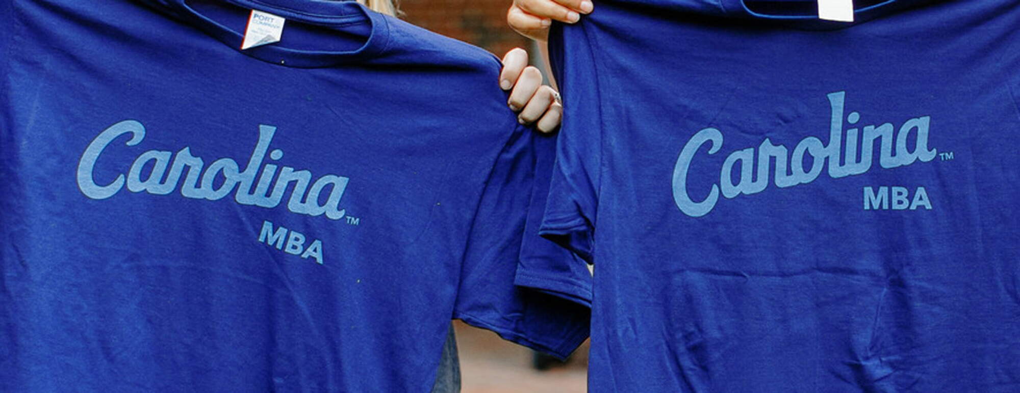 Two people are holding up t-shirts that say Carolina MBA