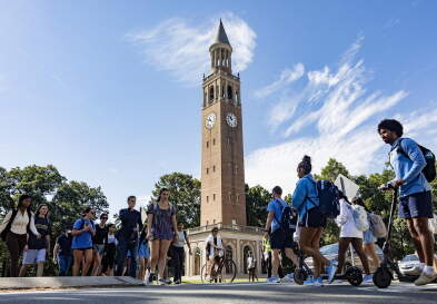 The Bell Tower against a Carolina blue sky with students crossing the street in front of it.