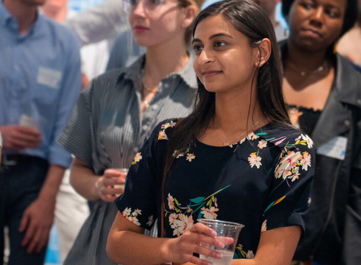 A woman stands in a crowd looking slightly off camera. She is holding a glass of water and is engaged in whatever is happening.