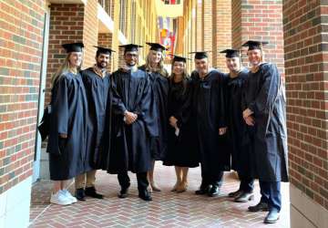 A group of eight graduates pose under the UNC Kenan-Flagler flags outside of McColl Building.