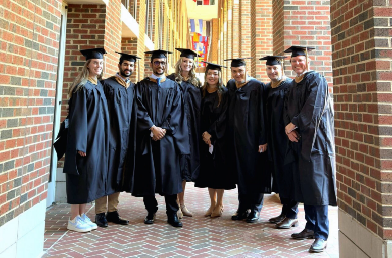 A group of eight graduates pose under the UNC Kenan-Flagler flags outside of McColl Building.