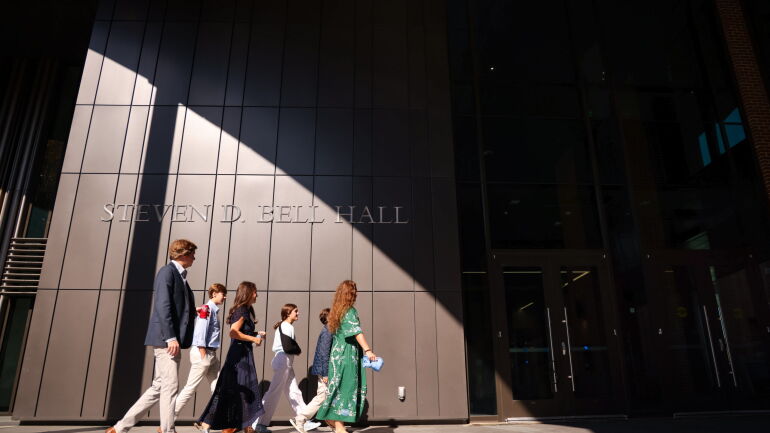 Attendees of the grand opening walk in the sunlight underneath the Steven D. Bell Hall lettering outside of the entrance.