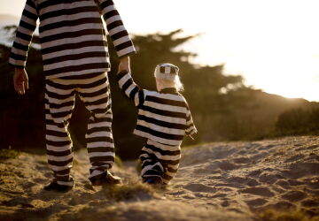Young boy walking hand in hand with his father while wearing a striped prisoner uniform on a beach at sunset.