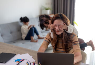 A woman sits on a laptop at home while a child covers their eyes.