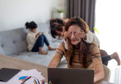 A woman sits on a laptop at home while a child covers their eyes.