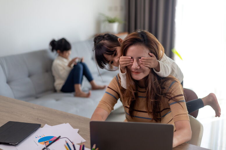 A woman sits on a laptop at home while a child covers their eyes.