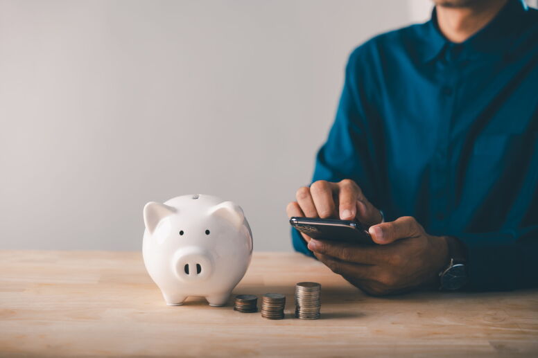 Person uses a smartphone next to a piggy bank.